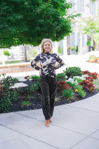 Full body front view of a smiling blonde woman posing outdoors with her hands on her hips, modeling a fitted black and beige abstract cow print long-sleeve top. The edgy, patterned shirt features a chic mock neckline and is tucked into high-waisted black corduroy pants. The textured wide-leg trousers are detailed with striking white contrast stitching outlining the front pockets, fly, and distinct knee seams. She completes the stylish, transitional fall look with brown leather square-toe boots and large gol