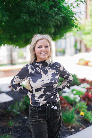 Front view of a smiling blonde woman posing outdoors with her hands on her hips, modeling a fitted black and beige abstract cow print long-sleeve top. The edgy, patterned shirt features a chic mock neckline and is tucked into high-waisted black corduroy pants. The textured trousers are detailed with striking white contrast stitching along the front pockets and waistband. She completes the transitional fall look with gold hoop earrings, set against a landscaped outdoor background.