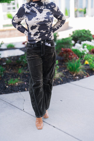 Front mid-body view of a woman posing outdoors with her hands on her hips, modeling a fitted long-sleeve mock neck top in a black and beige abstract cow print. The top is seamlessly tucked into high-waisted, dark corduroy pants featuring striking white contrast stitching along the front pockets, fly, and distinct horizontal knee seams. The relaxed, wide-leg textured trousers drape perfectly over tan leather square-toe boots, set against a landscaped background with lush greenery.