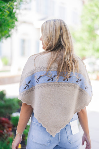 Back view of the beige knit poncho sweater showcasing its elegant, relaxed drape and distinctive pointed, scalloped hemline. The continuous light blue and brown patterned band spans across the shoulders, highlighting the soft, fine-knit texture. The poncho is styled effortlessly with light wash blue denim jeans featuring classic back pockets and a visible white Lovervet brand tag, completing a breezy, transitional fall outfit.