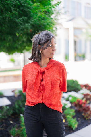 Woman looking to the side, modeling a vibrant red short-sleeve knit sweater featuring a button-up front and a statement tie-neck bow. The cozy, textured knit top is styled with black denim jeans featuring subtle rhinestone embellishments. Accessorized with classic gold hoop earrings and a claw clip, the outfit offers a chic, elevated everyday look set against an outdoor backdrop.