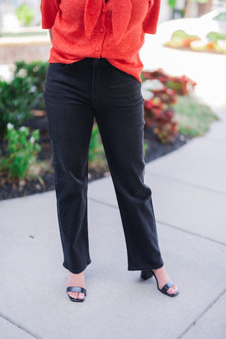 Lower body front view of a woman modeling black straight-leg denim jeans featuring sparkling rhinestone embellishments cascading down the outer side seams. The statement denim is styled for an elevated everyday look with a vibrant red textured short-sleeve sweater and black square-toe block heel sandals, photographed outdoors on a paved walkway.