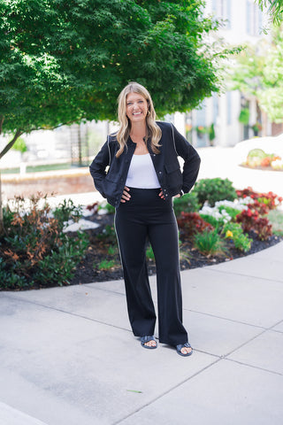 Full body front view of a smiling blonde woman posing outdoors with her hands on her hips, modeling a sporty-chic black two-piece athleisure set. The outfit features a cropped black bomber-style jacket with sleek white contrast piping along the shoulders and sleeves, dark snap-button closures, front flap patch pockets, and striped ribbed cuffs. Worn open over a fitted white scoop-neck top, the jacket pairs perfectly with matching high-waisted black wide-leg pants featuring a distinct white contrast side str