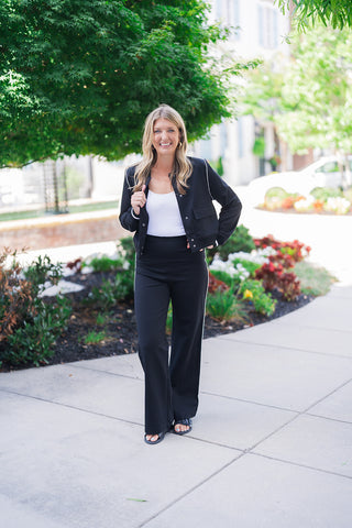 Full body front view of a smiling blonde woman posing outdoors, modeling a sporty-chic black two-piece athleisure set. The outfit features a cropped black bomber-style jacket with white piping details, front patch pockets, and striped ribbed cuffs, layered over a fitted white scoop neck top. She pairs the jacket with high-waisted black wide-leg pants featuring a distinct white contrast side stripe. The elevated casual look is styled with black buckle slide sandals and gold hoop earrings, set against a lands