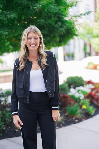 Front mid-body view of a smiling blonde woman posing outdoors, modeling a sporty-chic black bomber-style jacket featuring sleek white contrast piping along the shoulders and sleeves. The tailored jacket includes dark snap-button closures, front flap patch pockets, and striped ribbed cuffs, worn open over a fitted white scoop-neck top. She pairs the layer with matching high-waisted black pants that showcase a white side stripe, completing the elevated athleisure look with classic gold hoop earrings.