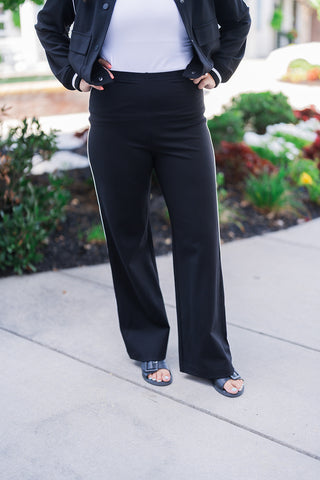 Front mid-body view highlighting the flattering fit and styling of the black matching set. The image showcases the smooth, high-waisted silhouette of the black wide-leg pants and the elongating effect of the white side stripe. The model poses with hands on her hips, wearing the black jacket open over a crisp white top to emphasize the comfortable, everyday athleisure aesthetic, completed with black slide sandals.