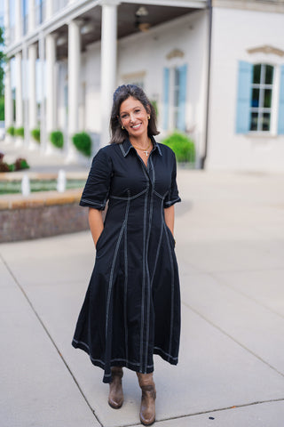 Woman smiling outdoors wearing a black button-down midi shirtdress featuring striking white contrast topstitching. The dress has a classic collared neckline, elbow-length sleeves, and functional side pockets. Styled for a chic, transitional look with brown leather heeled ankle boots and a gold cross necklace, standing on a paved walkway in front of a white building with columns and light blue shutters