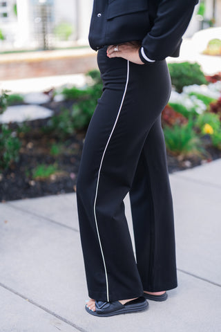 A detailed close-up side view focusing on the crisp, tailored accents of the black athleisure set. The image clearly highlights the continuous white piping running down the side seam of the pants, matching the sporty striped ribbed cuff of the jacket sleeve. The model rests a hand on her hip, showcasing the comfortable stretch fabric and sleek, retro-inspired sportswear design.