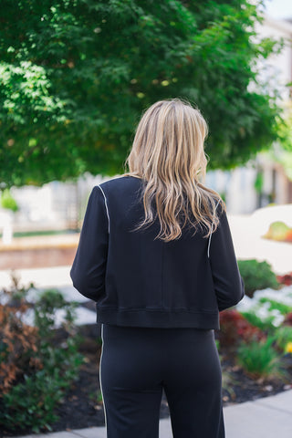 Upper body back view of the black bomber jacket highlighting its clean, structured silhouette and comfortable fit. The image showcases the striking white contrast piping detailing on the back of the sleeves and a thick, comfortable ribbed bottom hem. The sporty jacket rests cleanly at the waistband of matching black athleisure pants, emphasizing a polished yet relaxed everyday aesthetic set against a lush, landscaped background.