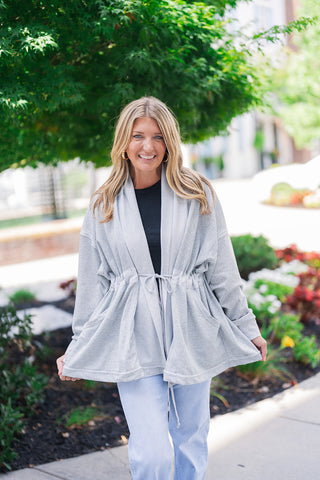 Front view of a smiling blonde woman posing outdoors, modeling a light grey open-front jacket made of a soft, sweatshirt-like material. The relaxed athleisure piece features wide lapels, drop shoulders, and a functional drawstring waist that ties in the front to create a flattering peplum silhouette. She styles the versatile, transitional layer over a simple black top and light-wash blue denim jeans, accessorizing with classic gold hoop earrings against a landscaped background.