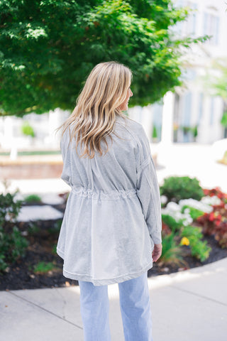 Back view of the light grey jacket highlighting its comfortable drape and unique structural details. The image showcases how the gathered drawstring waist beautifully cinches the back, creating a gentle flare at the hem. The cozy, french terry-style fabric features visible drop-shoulder seams and a relaxed fit, paired perfectly with light-wash blue jeans for an effortless, everyday look.