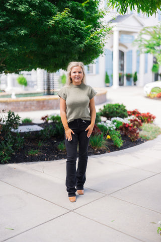 Full body front view of a smiling blonde woman posing outdoors, modeling a transitional fall outfit. She is wearing an olive green short-sleeve knit sweater that features a ribbed mock neckline, ribbed cap sleeves, and a soft, cozy texture. The top is styled with a casual front-tuck into a pair of black bootcut denim jeans. The elevated everyday look is completed with brown leather square-toe boots, gold hoop earrings, and layered delicate bracelets, set against a beautifully landscaped garden and a classic