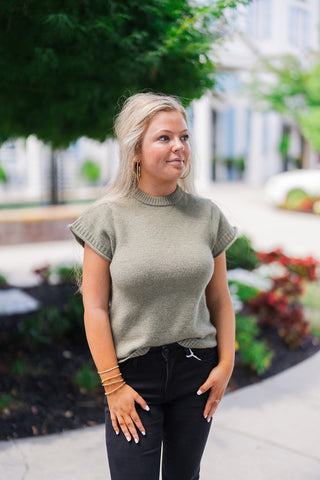 Front view of a blonde woman posing outdoors, modeling a cozy olive green short-sleeve knit sweater. The soft, textured top features a ribbed mock neckline, structured ribbed cap sleeves, and a comfortable relaxed fit. She styles the transitional seasonal piece with a subtle front tuck into black denim jeans. The everyday casual look is completed with gold hoop earrings and layered gold bracelets, set against a beautifully landscaped outdoor walkway and white building.