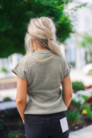Back view of the olive green knit sweater showcasing its comfortable drape and soft, finely knit texture. The image highlights the continuous ribbed detailing at the collar and sleeve openings. The sweater rests effortlessly over classic black denim jeans, which feature standard back patch pockets and a visible white Lovervet brand tag attached to the right pocket, emphasizing a trendy, effortless aesthetic.
