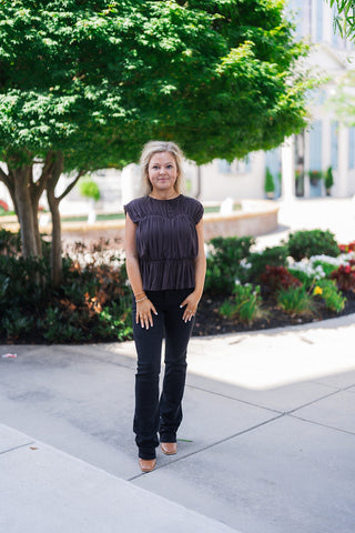 Woman standing outdoors wearing a dark charcoal pleated peplum blouse featuring a high round neckline and delicate cap sleeves. The top is styled casually but elegantly with black slightly flared jeans and brown square-toe shoes. The model is posing on a paved walkway bordered by manicured landscaping, trees, and a building with light blue shutters in the background.