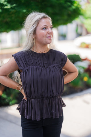 A portrait view of the dark charcoal blouse highlighting its intricate textural details, including all-over vertical pleating that gathers elegantly along an empire waistline before flaring out into a soft peplum hem. The cap sleeves create a subtle ruffle effect at the shoulders. The model has her hands on her hips and is accessorized with gold hoop earrings, beaded bracelets, and delicate necklaces.