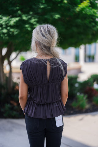 Back view of the dark charcoal pleated blouse showcasing a distinct keyhole cutout in the center upper back, secured at the neckline. The continuous vertical ruching and pleating extend smoothly down the back, flowing into the ruffled peplum hemline that rests over a pair of black denim jeans.
