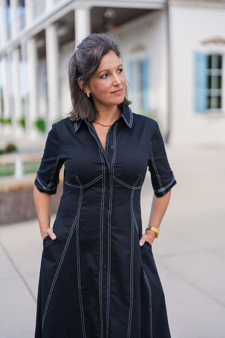 A portrait view of the black contrast stitch shirtdress highlighting the tailored, paneled bodice. The white topstitching creates architectural, corset-inspired lines that flatter the waistline. The dress features a crisp collar, dark button-front closure, and stitched cuffs on the short sleeves. The woman has her hands resting in the dress's side pockets, accessorized with a gold cross pendant, small hoop earrings, and a gold watch for a polished, everyday aesthetic.