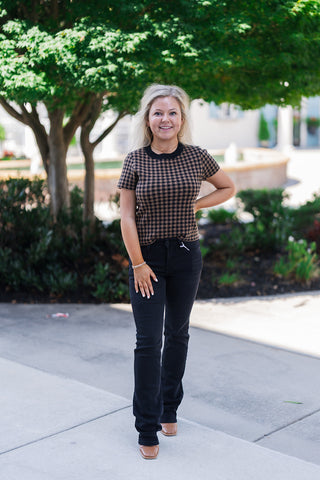 Full body front view of a smiling blonde woman posing outdoors, modeling a fitted brown and black gingham short-sleeve knit top. The retro-inspired sweater tee features a contrasting black ribbed crew neckline and matching ribbed hems. She pairs the patterned top with black denim jeans and brown square-toe shoes. The everyday casual look is accessorized with gold hoop earrings and a delicate gold necklace, set against a landscaped background with a green tree and fountain.
