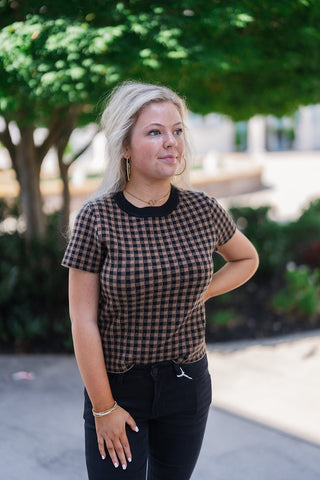 A portrait view of a woman looking off-camera with one hand on her hip, highlighting the texture and pattern of the brown and black checkered knit top. The close-up clearly showcases the soft, stretchy knit fabric, the classic black ribbed crew neckline, and the fitted short sleeves. She styles the top casually with black denim jeans and minimalist gold jewelry for a polished, transitional weather look.