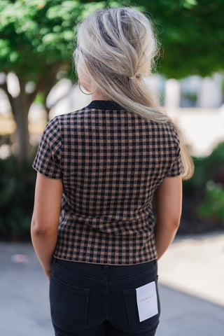 Back view of the brown and black gingham short-sleeve knit top, showcasing its tailored, comfortable fit across the shoulders and waist. The continuous checkered pattern is framed by the black ribbed collar. The top rests cleanly at the waistband of black denim jeans, which feature standard back patch pockets and a visible white Lovervet by Vervet brand tag attached to the right pocket.