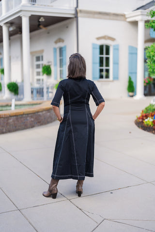 Back view of the black midi shirtdress showcasing the continuous white contrast stitching that forms elegant, elongating vertical panels from the shoulders down to the hem. The tailored fit of the bodice flows smoothly into a subtle A-line skirt, providing a comfortable yet structured silhouette. The look is grounded with brown leather ankle boots, emphasizing the dress's versatile movement and design.
