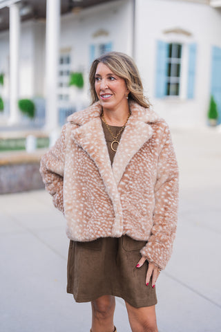 Full body front view of a woman looking off-camera and modeling a plush, fawn print faux fur jacket. The cropped, light brown coat features a distinctive white spotted pattern, an open front, and wide notch lapels. It is stylishly layered over an olive-brown faux-suede mini dress with front patch pockets. The chic, texture-rich fall outfit is accessorized with a gold necklace set against a classic white building with light blue shutters.