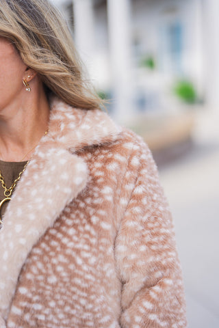 A detailed close-up view highlighting the ultra-soft, fuzzy texture of the fawn print faux fur jacket. The image focuses on the coat's structured wide notch collar and the delicate white spot pattern over the warm, light brown base. The neckline reveals the olive-brown faux-suede dress underneath, beautifully accented with gold chain necklace, and delicate crystal drop earrings.
