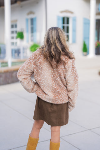 Back view of a woman with wavy hair modeling a plush, fawn print faux fur jacket. The cozy, light brown jacket features a distinct white spotted pattern and a relaxed, comfortable fit. The textured outerwear is styled for fall with a chocolate brown faux-suede mini skirt and tan leather knee-high boots, photographed outdoors against a classic white building with light blue shutters.