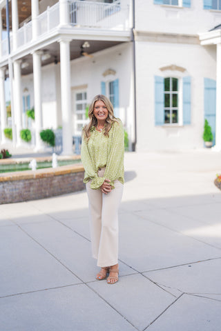 Full-length view of the Woodland Whisper Matcha Top styled with wide-leg cream trousers and nude braided heeled sandals. The top hangs loosely with a soft front tuck, emphasizing its fluid silhouette, relaxed fit, and feminine ruffle wrist detail against a bright architectural backdrop.