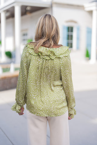 Back view of the Woodland Whisper Matcha Top highlighting the wide statement ruffle collar lining the neckline and subtle back gathers that create an effortless, relaxed drape. Features long blouson sleeves with ruffled elastic wrist cuffs over cream wide-leg pants.
