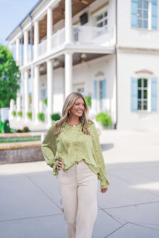 Woman posing outdoors in the Woodland Whisper Matcha Top, a lightweight satin-like blouse featuring a matcha green print with cream micro-abstract dots. The top showcases ruffled elastic cuffs, a split neckline with keyhole drawstring details, and a ruffled collar. Paired with cream wide-leg trousers and gold jewelry in front of an elegant white venue with light blue shutters.