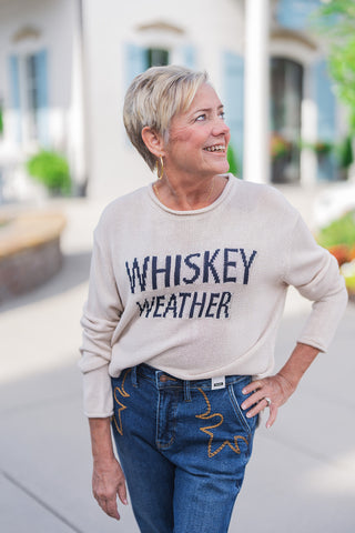 Front view of a smiling woman looking off-camera, modeling a lightweight beige knit sweater featuring the playful phrase "WHISKEY WEATHER" in contrasting dark navy block lettering across the chest. The cozy long-sleeve top features a relaxed fit and a casual rolled crew neckline. It is styled effortlessly with medium-wash blue denim jeans that showcase unique western-inspired star embroidery on the front pockets. She accessorizes the casual fall outfit with gold hoop earrings and a delicate gold necklace, s