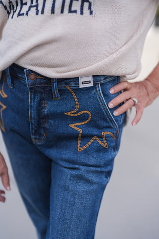 A detailed close-up view highlighting the front pocket and waistband of mid-rise, medium-wash blue denim jeans. The focal point is the striking western-inspired star embroidery, crafted with thick golden-brown contrast stitching across the front pocket. The image also clearly displays a bronze Judy Blue branded button at the closure and a white "MID RISE" sizing tag attached to the waistband. The jeans are styled with a soft beige knit sweater casually tucked in, with the model's hand resting on her hip.