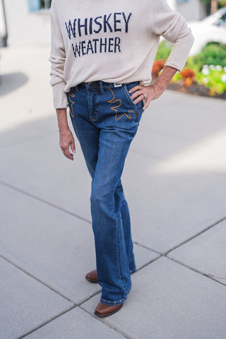 Angled lower body view highlighting the unique design details of the medium-wash blue jeans. The distinct gold-tone western star stitching on the front pockets stands out beautifully against the classic denim fabric. The comfortable, structured jeans feature standard belt loops and contrast stitching, draping perfectly over brown square-toe boots for a versatile, everyday fall look.
