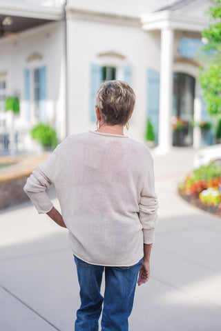 Back view of the beige knit sweater highlighting its comfortable, relaxed drape and lightweight, slightly sheer woven texture. The cozy long-sleeve pullover features subtle rolled hem detailing at the neckline, cuffs, and bottom edge. It rests easily over a pair of medium-wash blue denim jeans, emphasizing an effortless, trendy, and comfortable seasonal aesthetic.