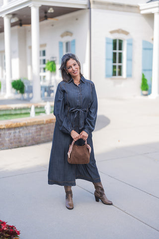 Full body front view of a smiling woman posing outdoors, modeling a chic dark wash denim midi dress. The versatile, utilitarian shirtdress features a classic collar, a functional button-front closure, long sleeves with gathered elastic cuffs, and a self-tie waist belt for a flattering, cinched silhouette. She accessorizes the transitional fall outfit with a brown suede handbag adorned with a patterned silk scarf and brown leather heeled boots, standing against a classic white building with light blue shutte