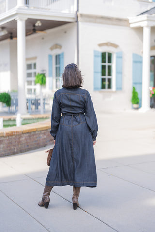 Full body back view of the dark wash denim midi dress showcasing its tailored, comfortable drape. The image highlights the structured back yoke, prominent gold contrast stitching along the seams, and a clean, flared midi-length hemline. The dress rests effortlessly over brown leather heeled boots, emphasizing a stylish, everyday seasonal aesthetic.