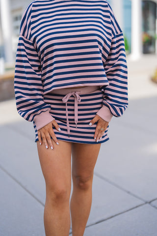 A close-up view highlighting the comfortable design details of the navy and pink horizontal striped two-piece set. The image focuses on the slightly cropped, relaxed fit of the long-sleeve pullover with its solid pink ribbed cuffs, paired perfectly with the high-waisted mini skirt featuring a functional solid pink drawstring tie and a ribbed hemline.