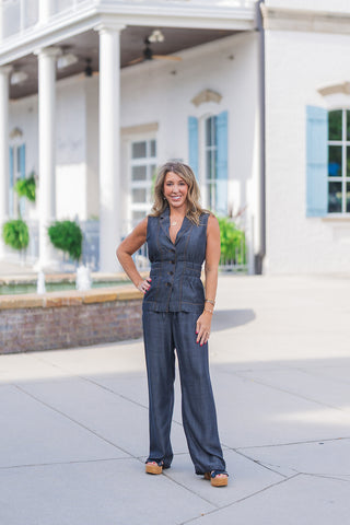 Full body front view of a smiling woman posing outdoors, modeling a matching dark wash chambray two-piece set. The outfit features a tailored sleeveless vest with a classic lapel collar, V-neckline, dark button-front closures, and a cinched elastic waist, paired beautifully with matching flowy wide-leg pants. Detailed with contrast gold stitching, the chic, transitional outfit is styled with black platform wedge sandals, stacked gold bracelets, a gold pendant necklace, and a red manicure, set against a clas