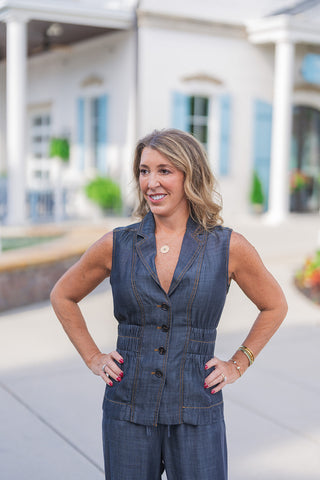 An upper body portrait view highlighting the tailored design of the dark wash chambray vest. The close-up showcases the vest's structured lapel collar, dark button front, and flattering smocked elastic waist that creates a subtle peplum silhouette. The prominent gold contrast stitching stands out beautifully against the dark denim-like fabric. She completes the elevated casual look with a gold compass pendant necklace, layered bracelets, and a bold red manicure, standing confidently with her hands on her hi