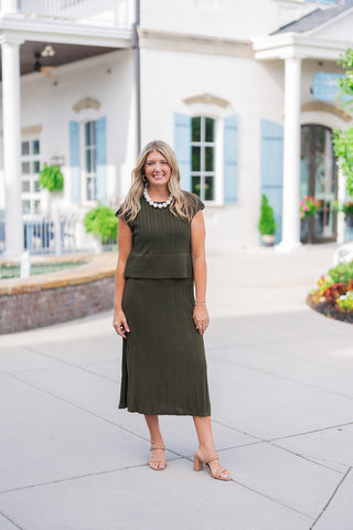 Full body front view of a smiling blonde woman posing outdoors, modeling a chic two-piece olive green ribbed knit set. The matching outfit features a cap-sleeve top with a classic crew neckline, vertical ribbing, and a contrasting horizontal ribbed hem. It is paired beautifully with a coordinating vertically ribbed midi skirt. The comfortable, transitional look is styled with a chunky white beaded statement necklace, gold hoop earrings, and tan braided double-strap block heels, set against a classic white b