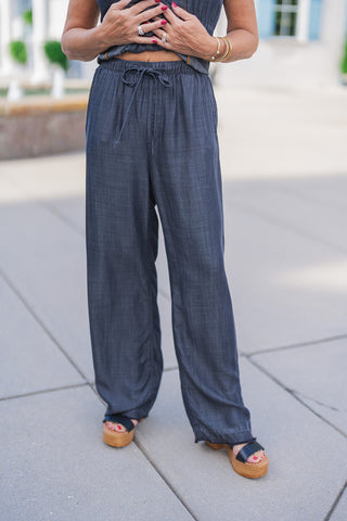 A close-up front view highlighting the comfortable, everyday design of the dark wash chambray wide-leg pants. The image focuses on the elastic gathered waistband with a functional drawstring tie and convenient side slip pockets. The woman's hands are resting near the waist, showing off a red manicure and silver rings, with the hem of the matching contrast-stitched vest visible just above. The relaxed-fit pants drape smoothly over black leather platform wedges.