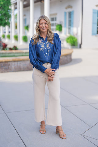 Full body front view of a smiling blonde woman posing outdoors, modeling a blue chambray long-sleeve blouse featuring intricate tan western-inspired embroidery on the chest. The button-up top is designed with voluminous balloon sleeves and wide, textured cuffs. The elevated casual top is styled with high-waisted beige wide-leg pants, tan woven block-heel sandals, and gold jewelry, set against a classic white building with light blue shutters.
