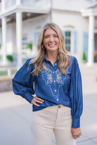 A portrait view highlighting the rich detailing of the blue chambray button-down blouse. The close-up showcases the striking tan symmetrical embroidery across the bodice, a classic collar, and light-colored buttons. The image captures the structural detail of the gathered shoulders and full balloon sleeves leading into elongated, fitted cuffs. The shirt is casually tucked into beige pants with visible front patch pockets, accessorized with gold hoop earrings and layered necklaces.