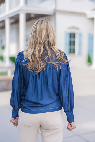 Back view of the blue chambray blouse showcasing its relaxed, airy silhouette. The design features a structured back yoke with subtle contrast stitching and gentle gathering beneath it, allowing the soft denim-like fabric to drape elegantly. The voluminous sleeves and long, tailored cuffs stand out against the smooth fit of the beige wide-leg pants, offering a comfortable yet polished everyday look.