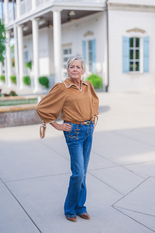 Woman posing outdoors wearing the Sweet Saddle Suede Blouse, a warm camel-tan faux suede top with contrast cream blanket-stitching along the collared V-neckline and tie-cuffs. Styled with medium-wash bootcut jeans featuring Western-embroidered pockets, a tan leather belt, and brown leather boots in front of a white building with light blue shutters.