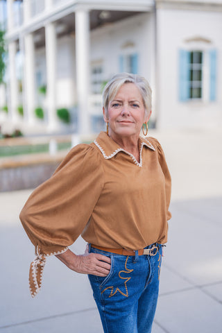 Upper-body view of the Sweet Saddle Suede Blouse highlighting the open split V-neck, structured collar trimmed with cream whipstitch embroidery, and dramatic volume long sleeves with self-tie wrist cuffs. Accessorized with thick gold hoop earrings.