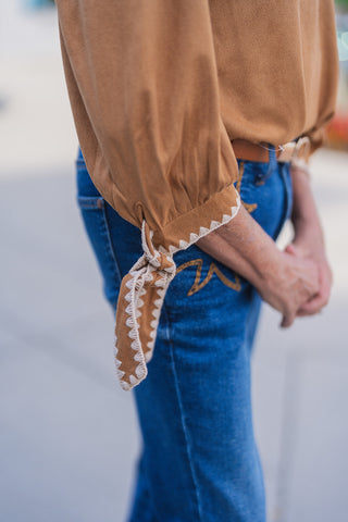 Detail shot focusing on the sleeve of the Sweet Saddle Suede Blouse, demonstrating the rich, soft faux-suede fabric texture, gathers at the wrist, and the intricate cream geometric blanket-stitch trim along the cuff tie closure.