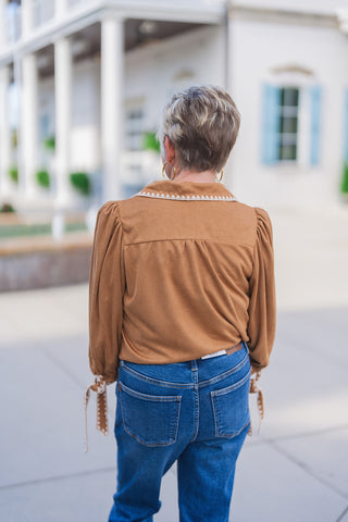 Back view of the Sweet Saddle Suede Blouse showing the whipstitched collar edge, back horizontal yoke seam with subtle gathering, and gathered puff shoulders. Paired with high-waisted blue jeans outdoors.